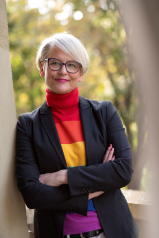 Bild von Sabine Mauer. Sie hat kurze blonde Haare, trägt eine schwarze Brille sowie einen Rollkragenpulli in Regenbogenfarben.